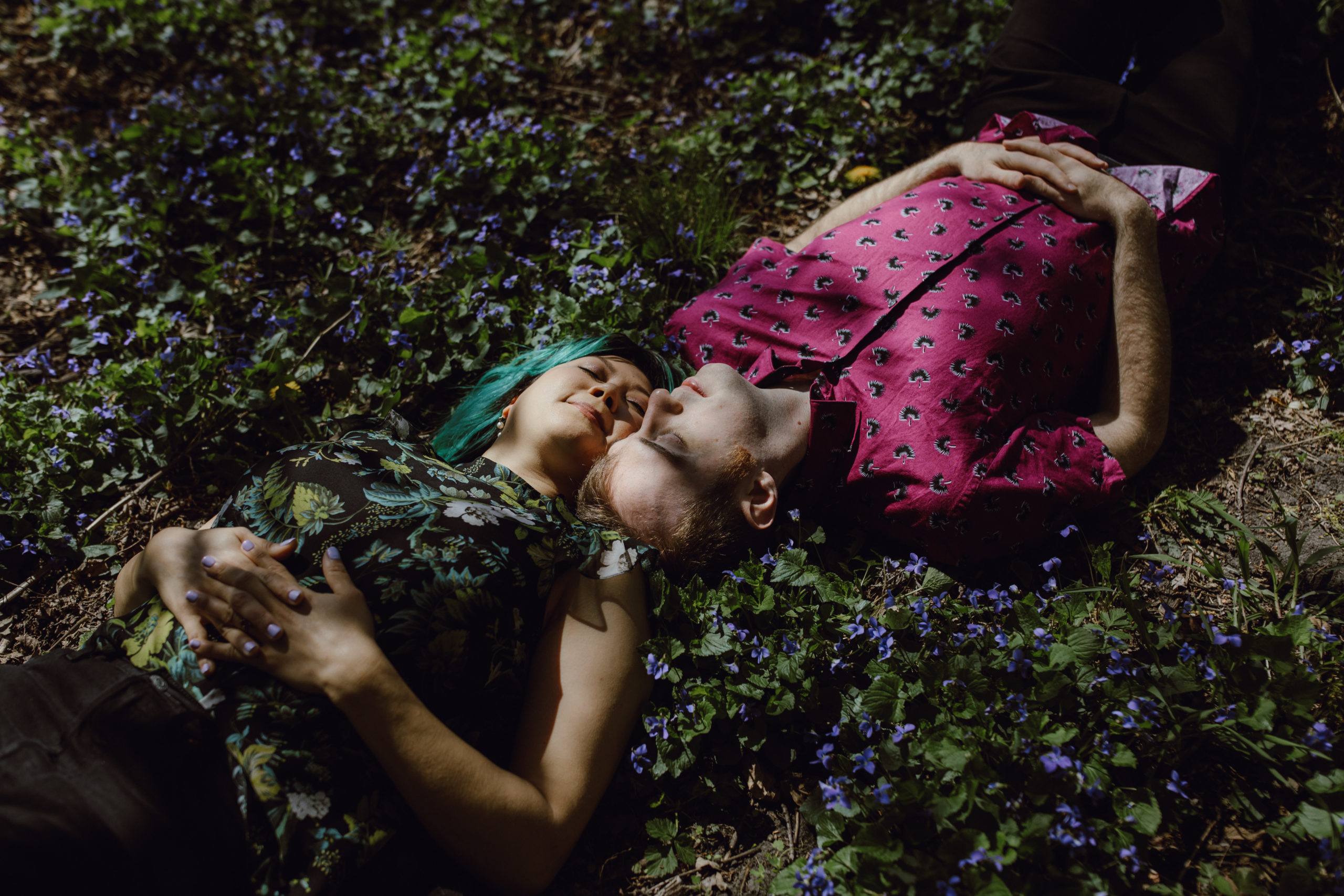 Couple laying on the ground surrounded by purple flowers with their eyes closed.